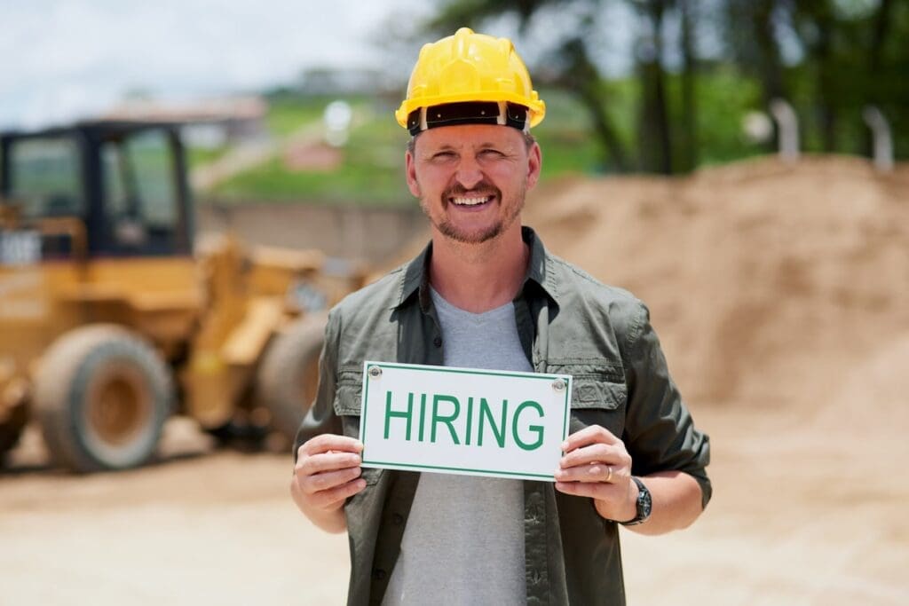 Online Marketing Strategies for Recruitment - Man in construction hat in front of equipment holding sign that says "Hiring"
